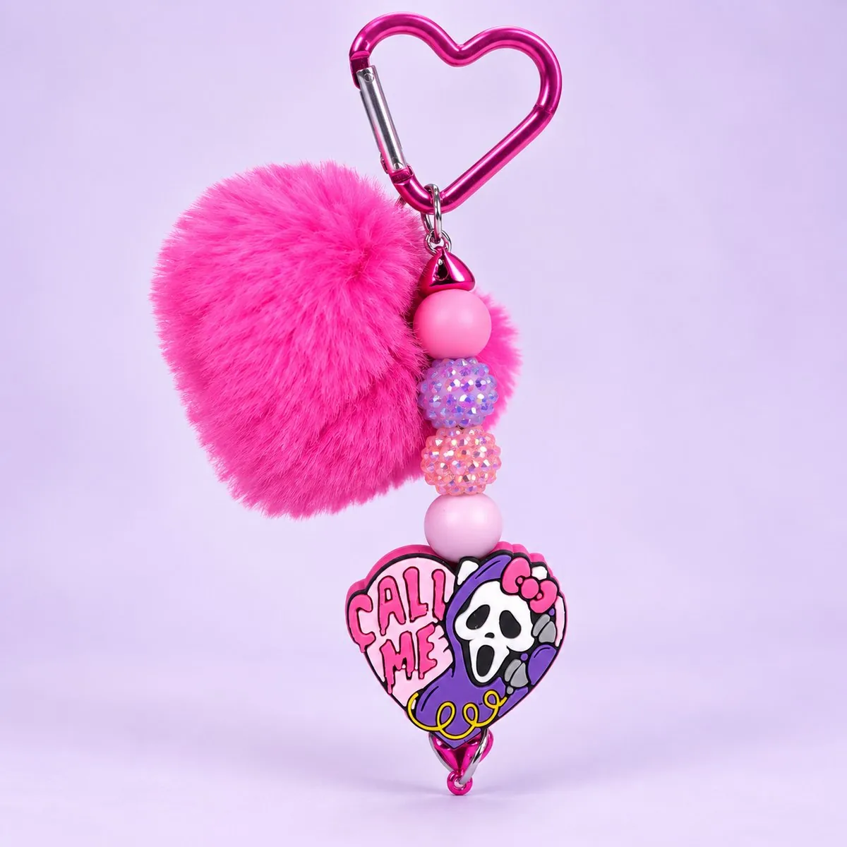 Call Me Ghost Heart Pom Charm on a pale lavender backdrop — a pink metal heart-shaped carabiner, a fluffy hot-pink heart pom-pom, pink, lilac and glitter beads, and a heart-shaped rubber charm reading 'CALL ME' with a white-masked ghost and a white bow cat