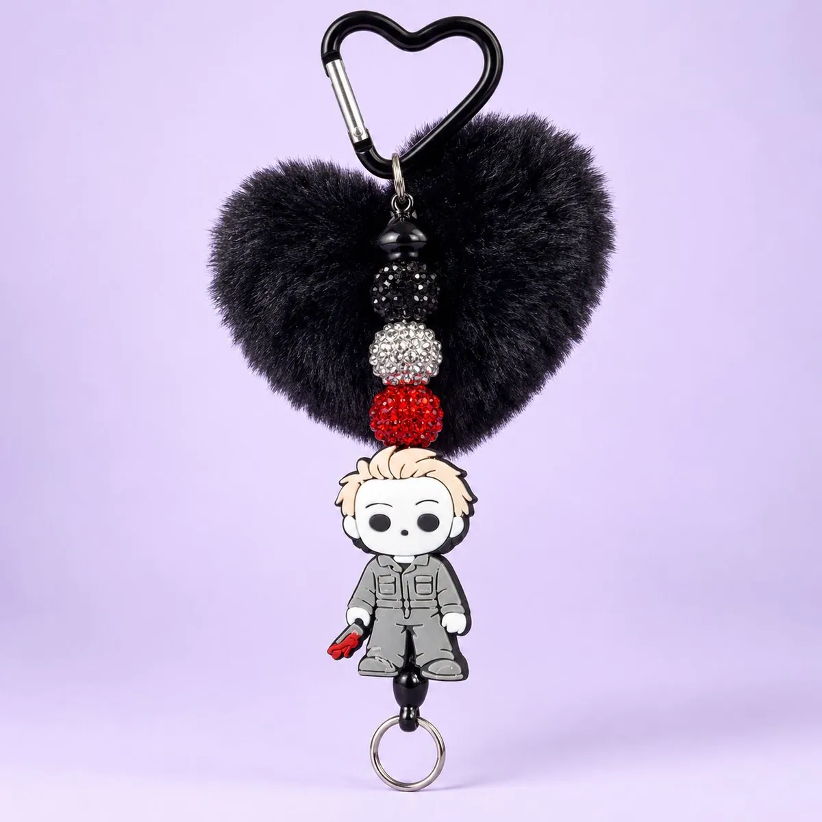 Masked Slasher Heart Pom Charm on a pale lavender backdrop — a black metal heart carabiner, a fluffy black heart pom-pom, black, silver and red rhinestone beads, and a rubber charm of a pale-masked man in a grey jumpsuit, with a silver split ring at the bottom