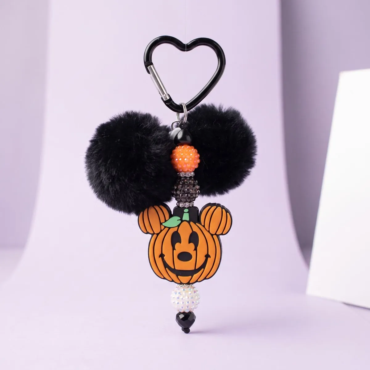 Pumpkin Mouse Charm hanging from its black heart-shaped carabiner — two black faux-fur poms above a carved pumpkin charm with round pumpkin ears and a green leaf, strung with orange and smoke rhinestone beads and a white iridescent bead