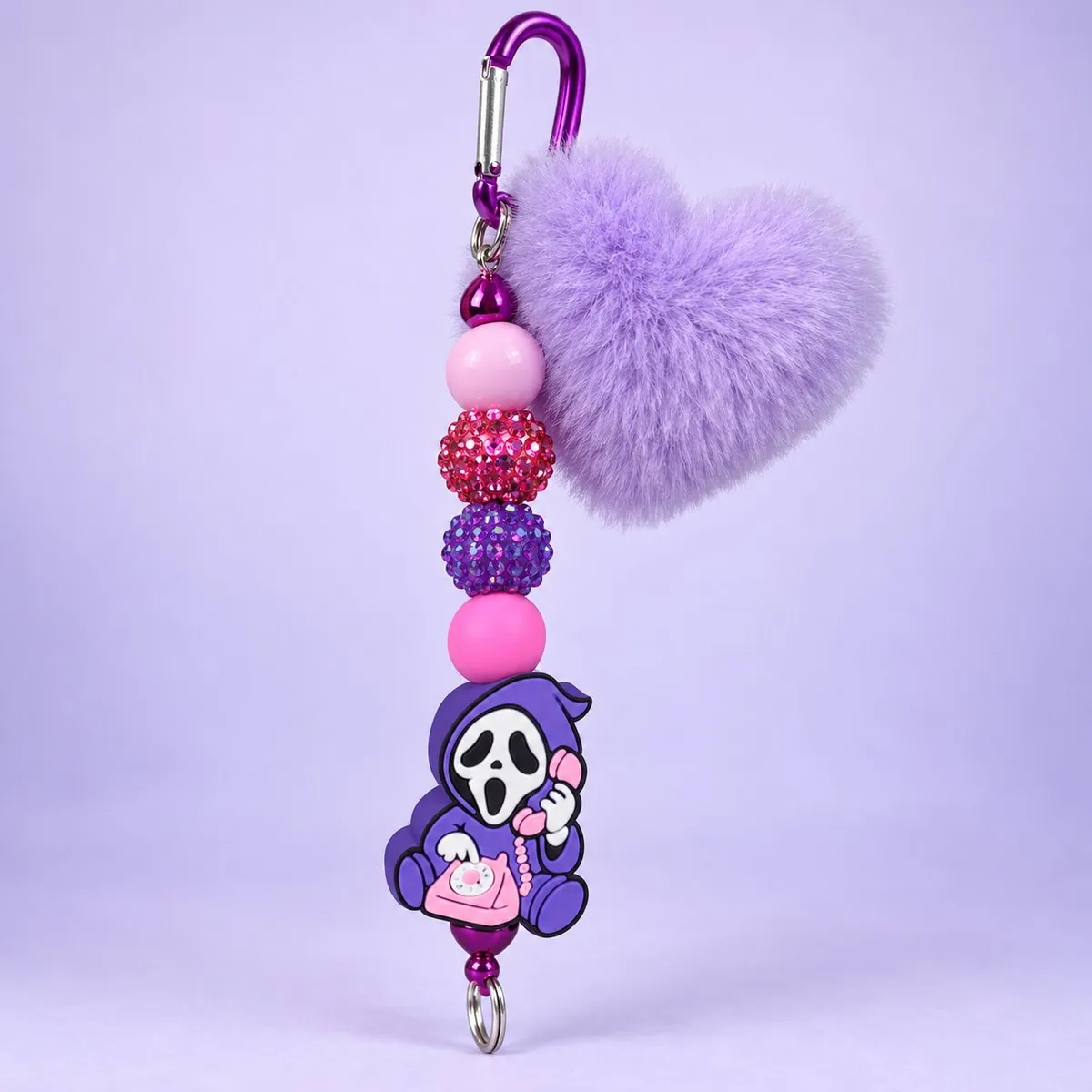 Purple Phone Ghost Heart Pom Charm on a pale lavender backdrop — keychain with the purple carabiner, a fluffy lilac heart pom-pom, pink, red and purple rhinestone beads, and a rubber charm of a purple-hooded white-masked ghost holding a pink telephone