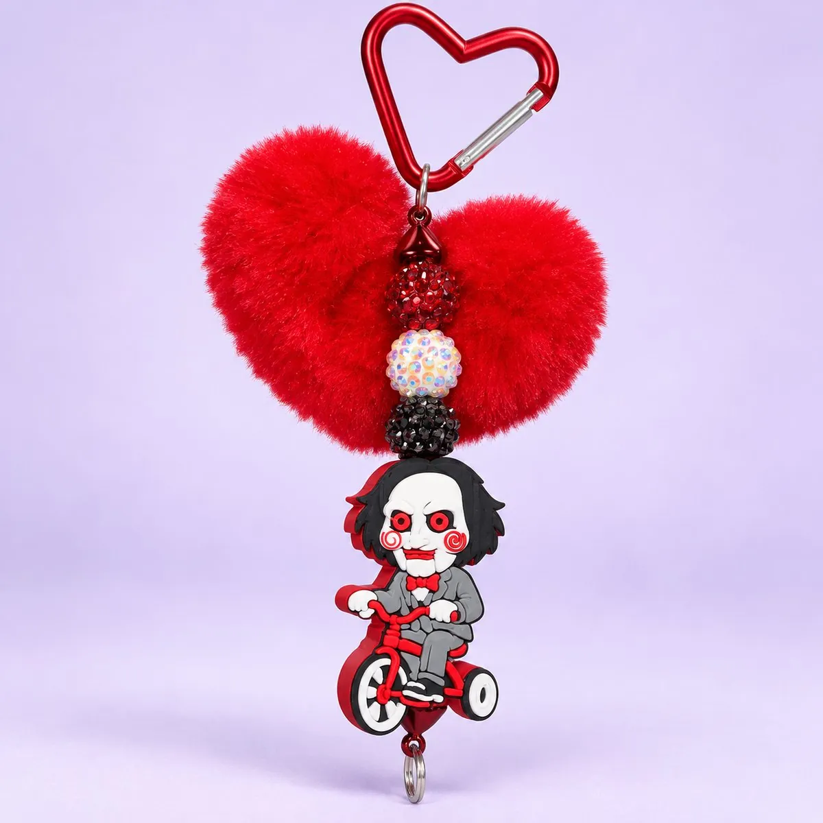 Tricycle Doll Heart Pom Charm on a pale lavender backdrop — keychain with the red metal heart carabiner, a fluffy red heart pom-pom, red and pastel rhinestone beads, and a rubber charm of a puppet doll with a white face and red spiral cheeks riding a red tricycle