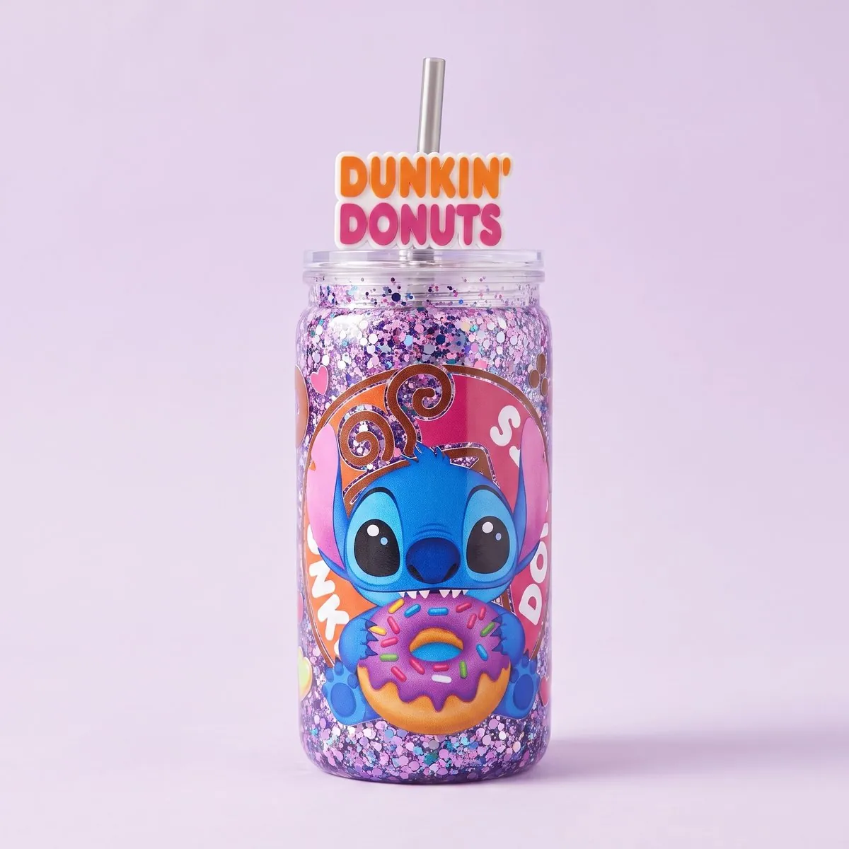 Blue Buddy Snow Globe Cup standing upright — a can-shaped tumbler packed with lilac and pink glitter behind a printed panel of a big-eared blue creature holding a sprinkled donut, with a clear lid, a metal straw and a lettered rubber straw topper