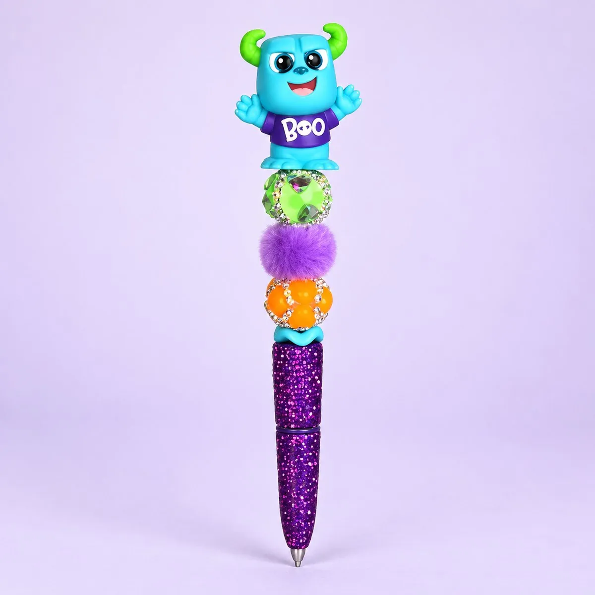 Boo Monster Beaded Pen on a pale lavender backdrop — beaded pen with a fluffy blue monster topper wearing a purple 'BOO' shirt, green, purple fluffy and orange beads, and a purple glitter barrel