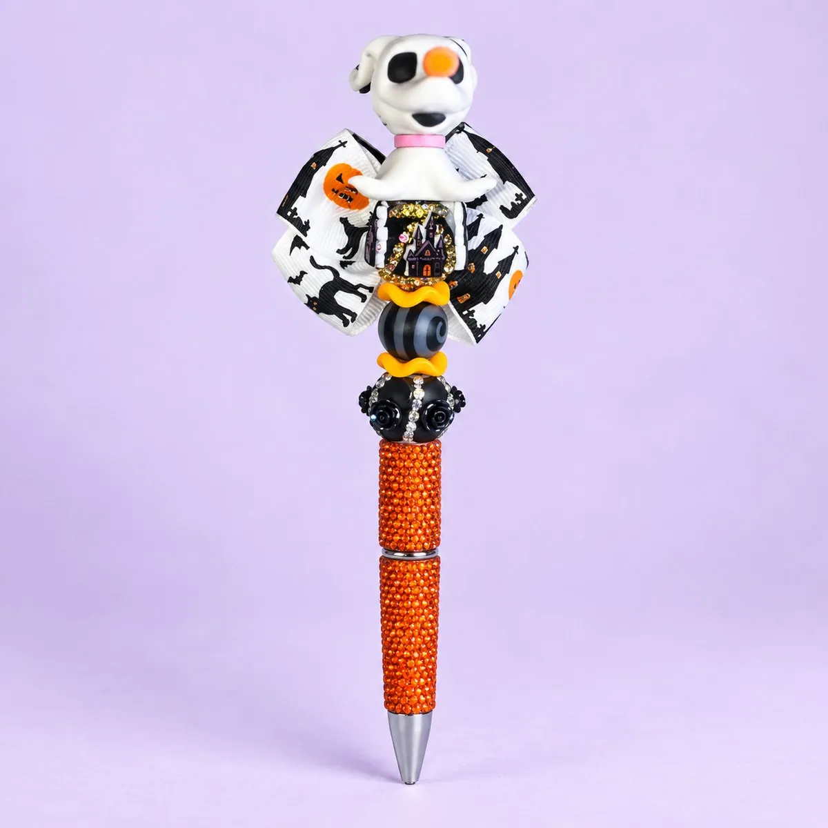 Ghost Pup Beaded Pen on a pale lavender backdrop — beaded pen with a white ghost dog topper, a black-and-white bat-print ribbon bow, orange and black beads, and an orange glitter barrel