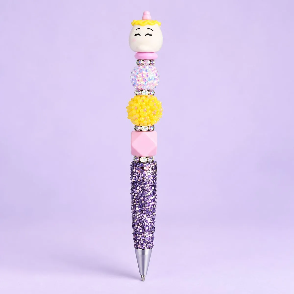 Pudding Pup Beaded Pen on a pale lavender backdrop — beaded pen with a round cream pudding-dog topper with a tiny crown, pastel, yellow and pink beads, and a lavender glitter barrel