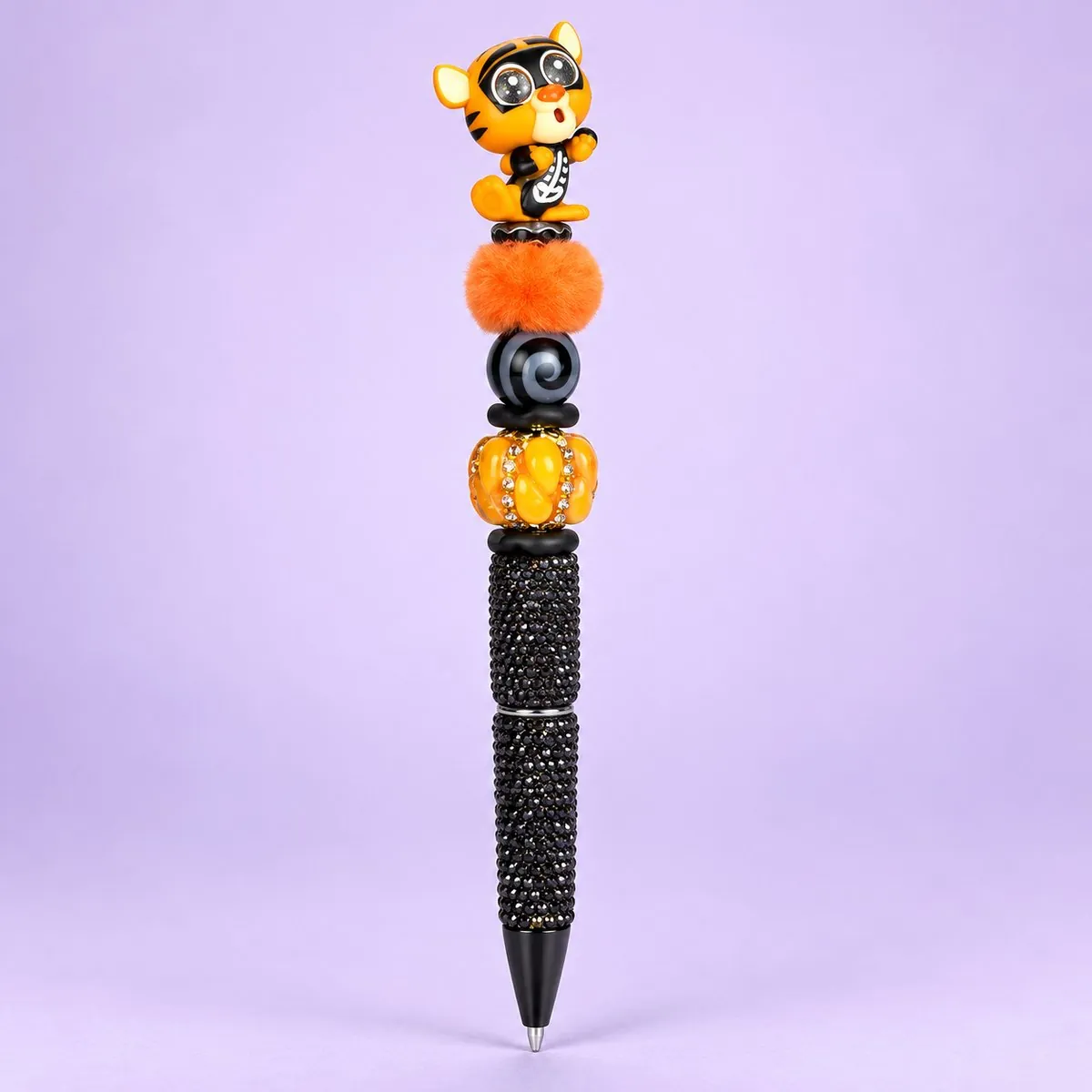 Skeleton Tiger Beaded Pen on a pale lavender backdrop — beaded pen with an orange striped tiger in a skeleton costume topper, orange, black swirl and pumpkin beads, and a black glitter barrel