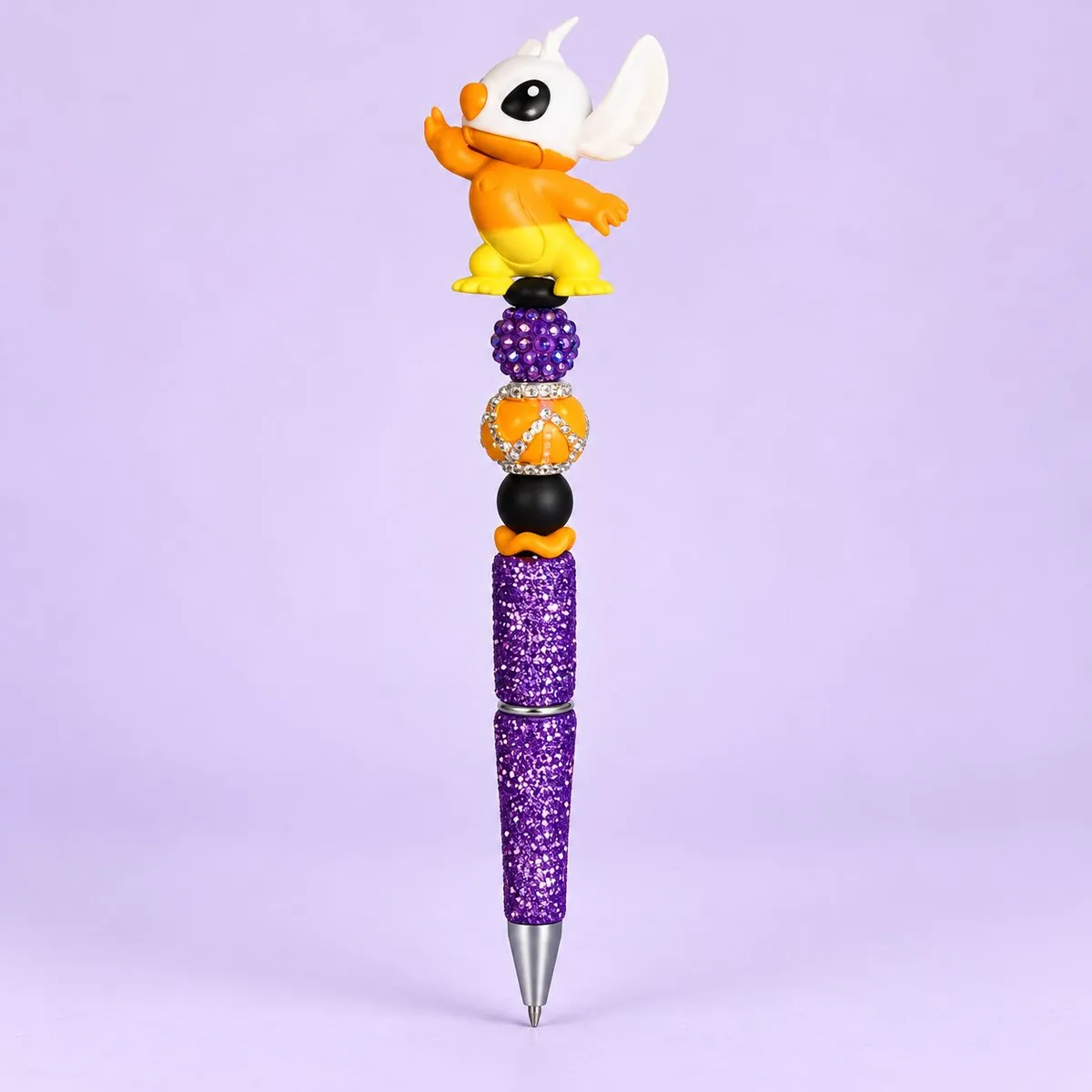 Spooky Alien Pup Beaded Pen on a pale lavender backdrop — beaded pen with a white-and-orange alien creature topper with big ears in a costume, purple, orange and black beads, and a purple glitter barrel