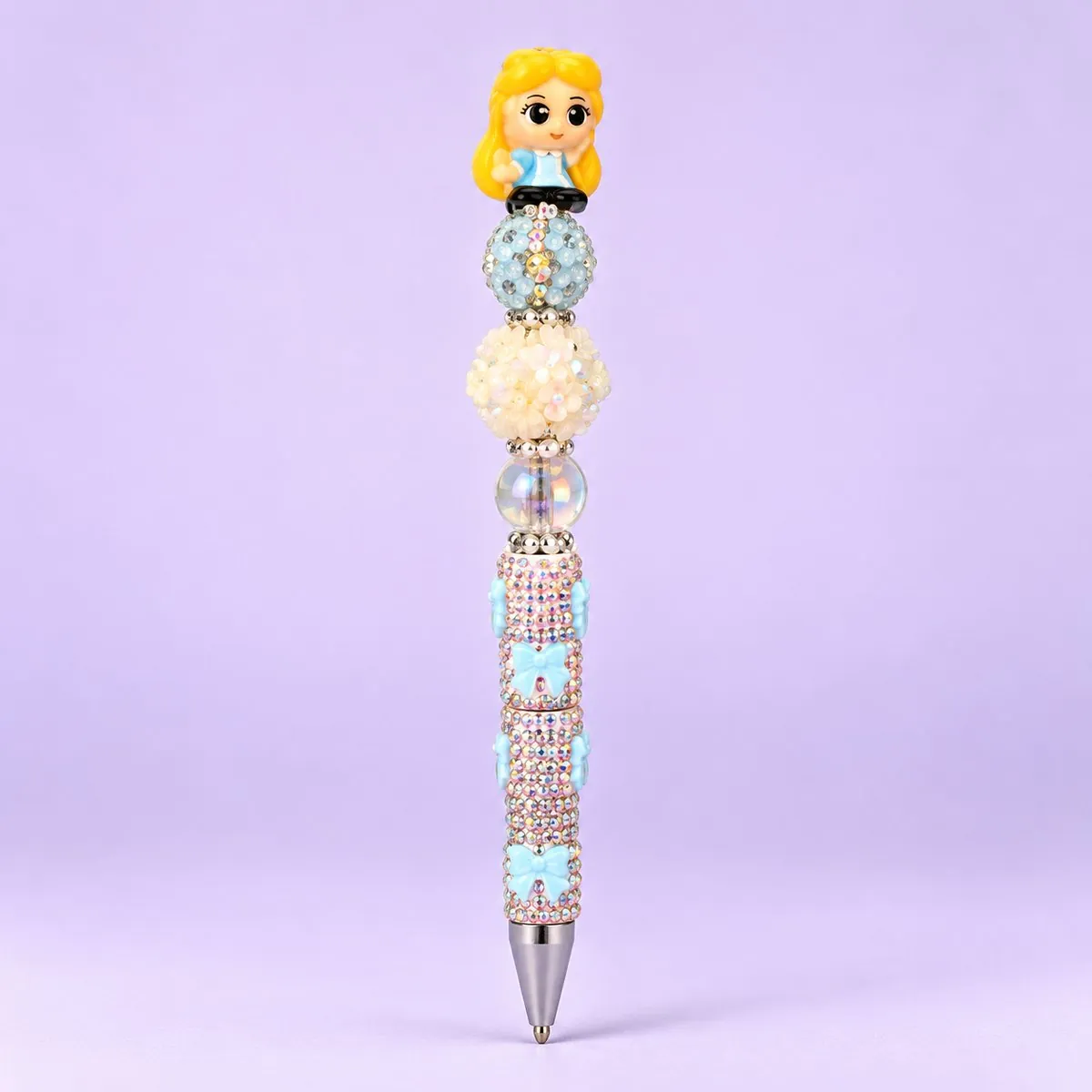 Wonderland Girl Beaded Pen on a pale lavender backdrop — beaded pen with a blonde girl in a blue dress topper, pale blue, cream flower and clear beads, and a pastel rhinestone barrel with blue bows